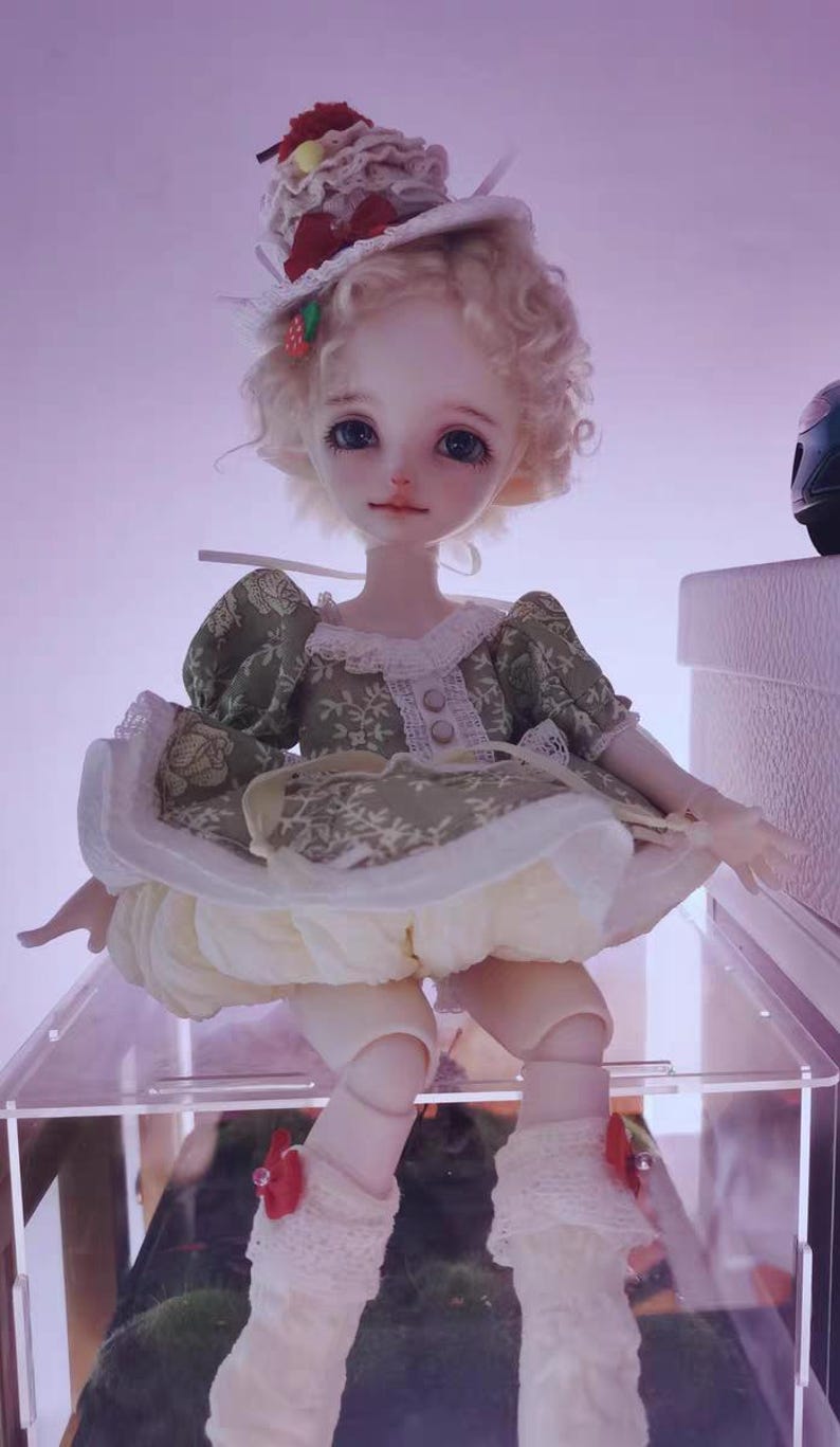 May include: A detailed doll with a whimsical design. The doll has a fair complexion, blonde curly hair, and is wearing a green dress with a white lace collar and a cream-colored skirt. A decorative hat with red accents tops the doll's head.