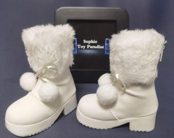 May include: A pair of white faux fur boots with pom poms. The boots have a zipper on the side and a platform heel. The boots are on a gray surface. The text "Sophie Toy Paradise" is visible in the background.
