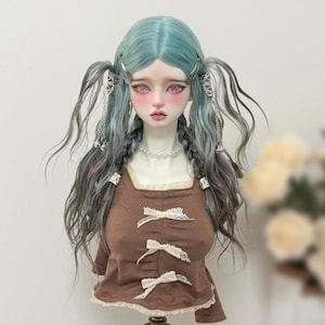 May include: A ball-jointed doll bust featuring long, wavy, teal and grey hair styled in pigtails with silver hair clips and dangling charms. The doll wears a brown top with three white lace bows down the front, a lace hem, and a pearl necklace.