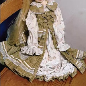 May include: A doll wearing a green and white dress with a lace trim. The dress has a floral pattern and a white collar. The doll has long blonde hair.