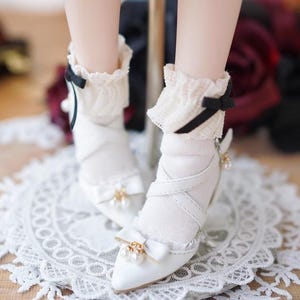 May include: White doll shoes with a pointed toe, adorned with small bows, pearls, and straps. The shoes are paired with white socks featuring a ruffled trim and black bows. The shoes are on a white lace doily.