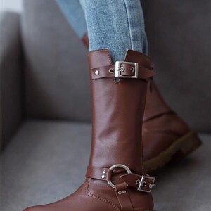 May include: Brown leather knee-high boots with silver buckles and a decorative ring on the strap. The boots have a chunky heel and a rounded toe.
