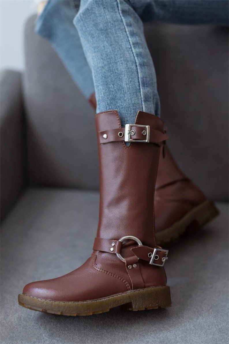 May include: Brown leather knee-high boots with silver buckles and a decorative ring on the strap. The boots have a chunky heel and a rounded toe.