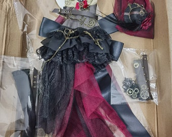 Op de afbeelding: Een steampunk-thema poppenoutfit met een zwarte en bordeauxrode tule rok, een bruin korset met gouden accenten en een zwart lint. De outfit bevat ook een zwarte hoed met een gouden tandwiel en een bordeauxrode sluier.