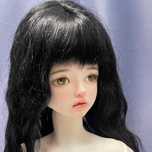 May include: A ball-jointed doll head featuring long, wavy black hair with bangs, pale skin, and large, detailed hazel-green eyes. The doll has a soft, neutral facial expression with subtle pink-toned lips and defined, realistic facial features.