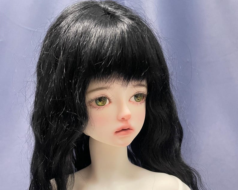 May include: A ball-jointed doll head featuring long, wavy black hair with bangs, pale skin, and large, detailed hazel-green eyes. The doll has a soft, neutral facial expression with subtle pink-toned lips and defined, realistic facial features.