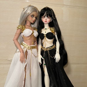 May include: Two dolls wearing elaborate gold and jeweled costumes. One doll has white hair and a white skirt, the other has black hair and a black skirt.