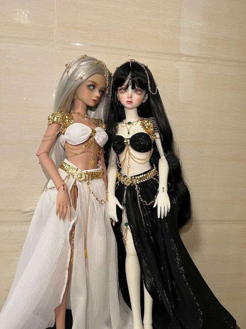May include: Two dolls wearing elaborate gold and jeweled costumes. One doll has white hair and a white skirt, the other has black hair and a black skirt.