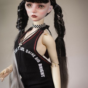 Puede incluir: Una mu&ntilde;eca con el pelo largo trenzado negro y gris que lleva un top negro sin mangas con ribetes rojos y el texto "The Stars Shine And Bright". La mu&ntilde;eca lleva un collar de gargantilla a cuadros blanco y negro y un collar de cruz plateado.