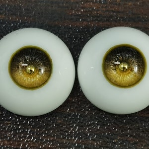 Puede incluir: Dos ojos de mu&ntilde;eca marrones y dorados con un acento dorado. Los ojos est&aacute;n colocados en una base de pl&aacute;stico blanco.