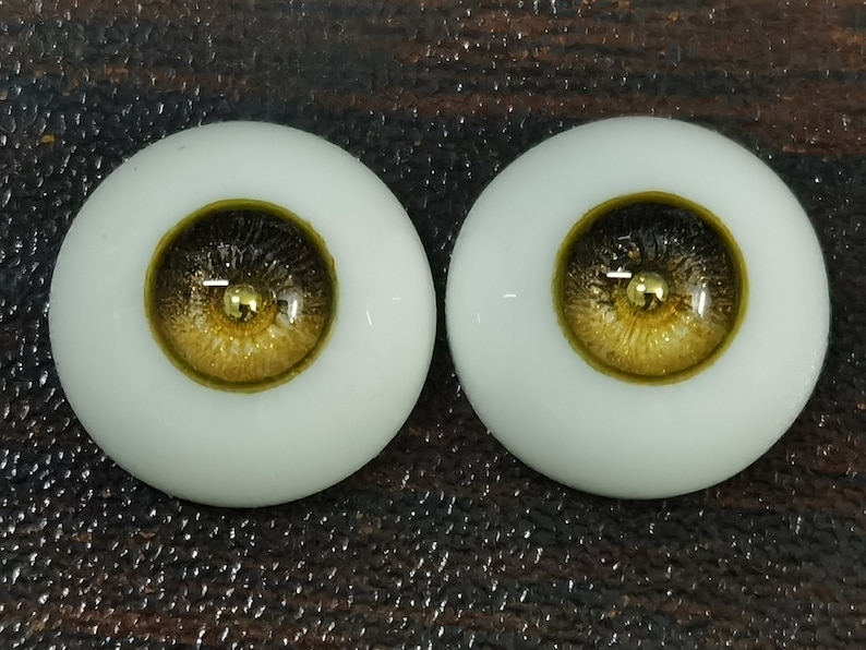 Puede incluir: Dos ojos de mu&ntilde;eca marrones y dorados con un acento dorado. Los ojos est&aacute;n colocados en una base de pl&aacute;stico blanco.