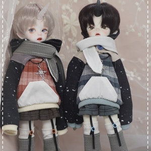 May include: Two dolls wearing plaid and patterned hooded sweatshirts with a white scarf and black and white checkered leggings. The dolls have white skin, black hair, and a horn on their head.