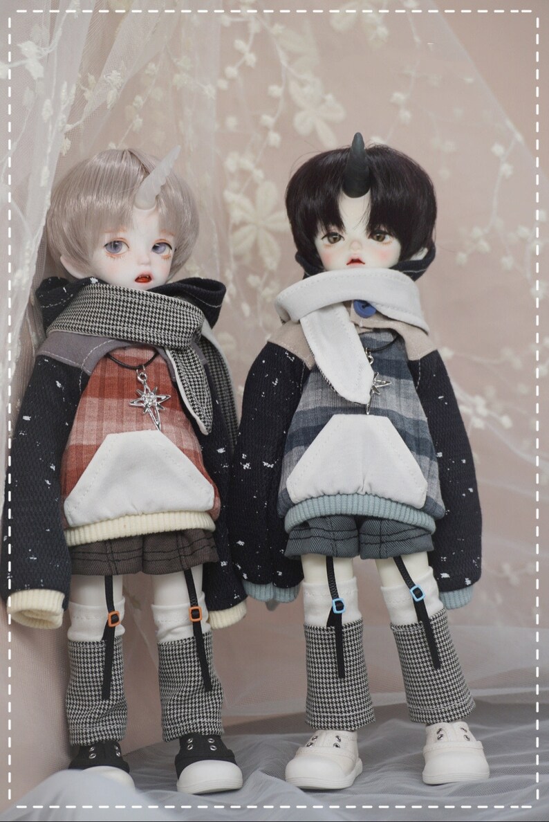 May include: Two dolls wearing plaid and patterned hooded sweatshirts with a white scarf and black and white checkered leggings. The dolls have white skin, black hair, and a horn on their head.