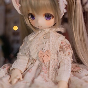 May include: A doll with long blonde hair styled in pigtails, wearing a white lace dress with pink floral accents and a pink bow. The doll has purple eyes and a delicate facial expression.