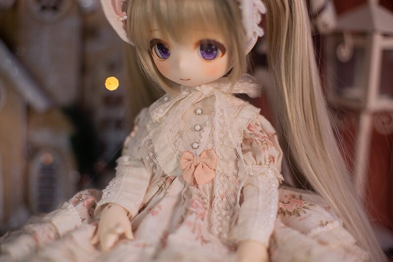 May include: A doll with long blonde hair styled in pigtails, wearing a white lace dress with pink floral accents and a pink bow. The doll has purple eyes and a delicate facial expression.