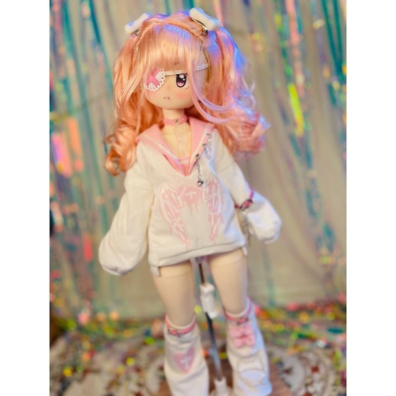 K&ouml;nnte beinhalten: Eine rosa und wei&szlig;e Puppe mit rosa Haaren, die ein wei&szlig;es Hoodie mit rosa Engelsfl&uuml;geln, eine rosa Halskette und wei&szlig;e Socken mit rosa Akzenten tr&auml;gt.