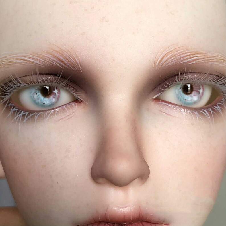May include: Close-up of a doll's face with white eyelashes and blue eyes with a pink and brown design.