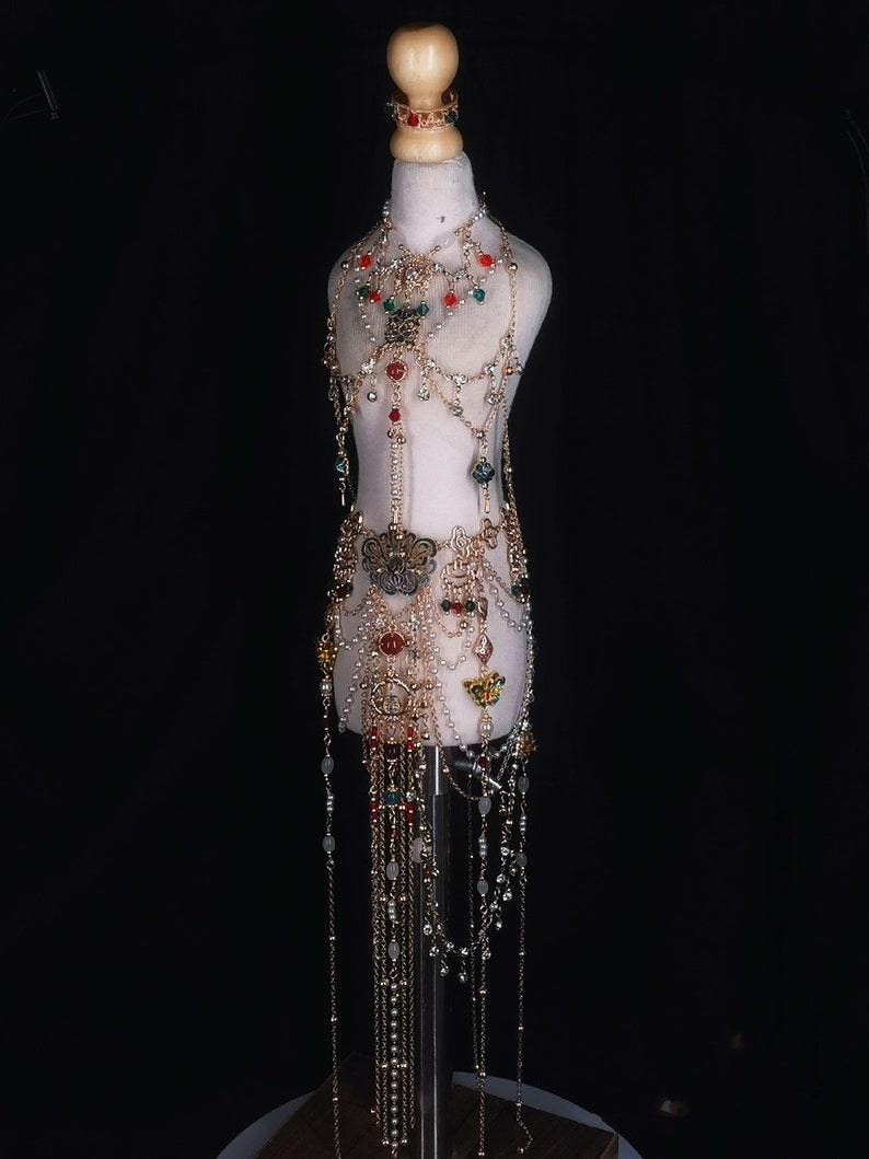 May include: A gold-toned body chain with red, green, and clear beads. The chain has a butterfly design and is layered with multiple strands of beads and chains.