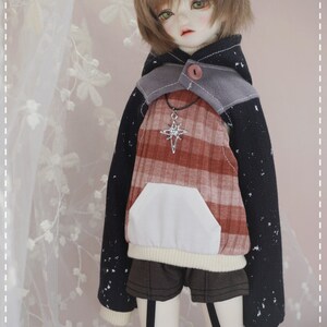 May include: A doll wearing a black and red plaid hoodie with a white pocket and a silver star necklace. The hoodie has a gray hood with a button. The doll is wearing black shorts and white socks with black suspenders.