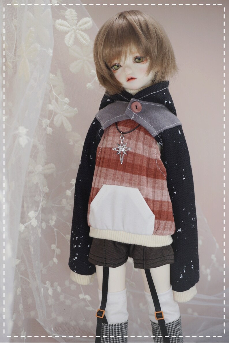 May include: A doll wearing a black and red plaid hoodie with a white pocket and a silver star necklace. The hoodie has a gray hood with a button. The doll is wearing black shorts and white socks with black suspenders.