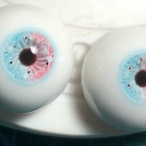 May include: Two realistic-looking glass eyes with blue, pink, and brown irises. The eyes are on a white surface.