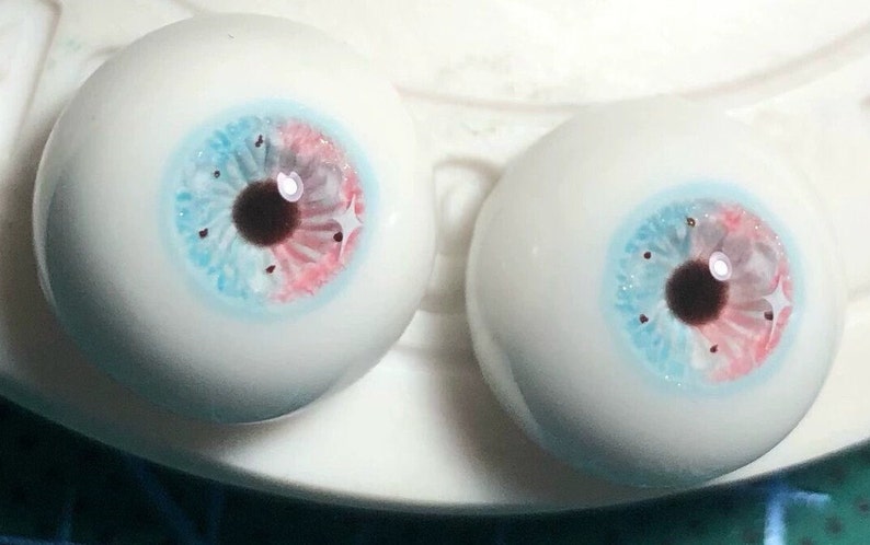 May include: Two realistic-looking glass eyes with blue, pink, and brown irises. The eyes are on a white surface.