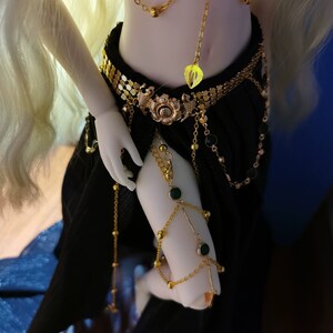 May include: A doll wearing a black skirt and gold jewelry with green gemstones. The jewelry includes a belt, a necklace, and bracelets.