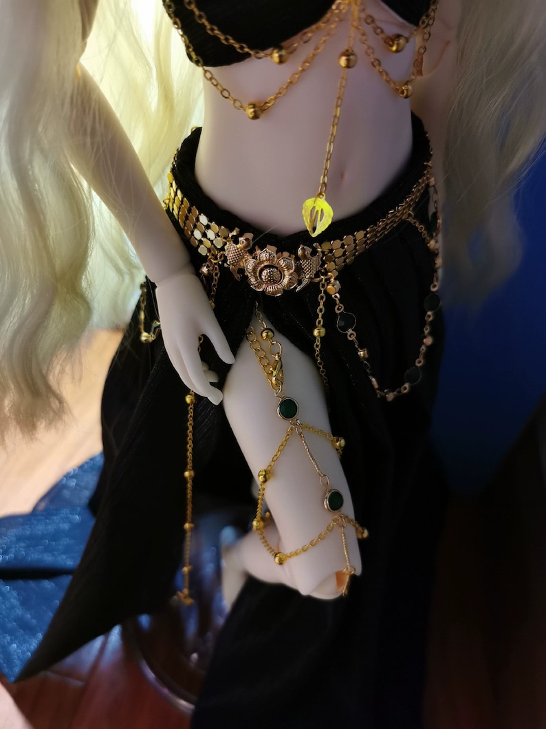 May include: A doll wearing a black skirt and gold jewelry with green gemstones. The jewelry includes a belt, a necklace, and bracelets.