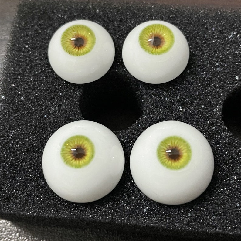 May include: Four realistic-looking glass eyes with white sclera, green irises, and brown pupils. The eyes are arranged in a square pattern on a black background.