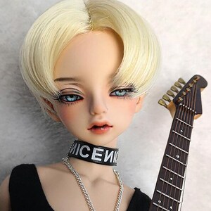 May include: A doll with short, straight, blonde hair wearing a black choker with the word "NICE" written on it. The doll is wearing a black tank top and has blue eyes. The doll is holding a guitar.