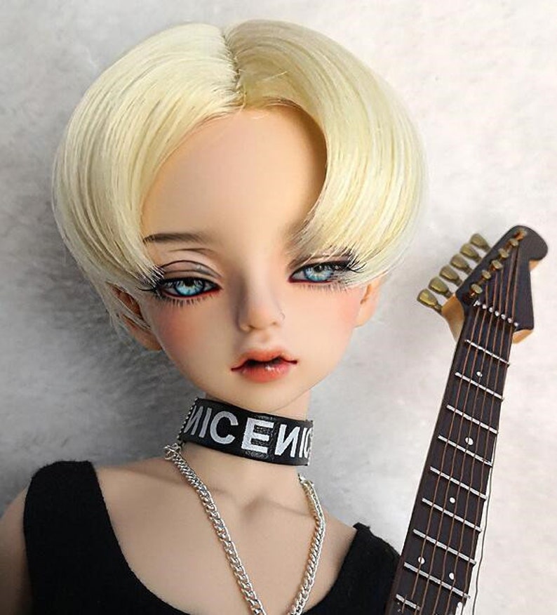 May include: A doll with short, straight, blonde hair wearing a black choker with the word "NICE" written on it. The doll is wearing a black tank top and has blue eyes. The doll is holding a guitar.