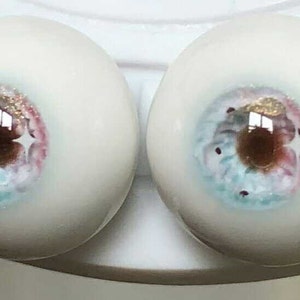 May include: Two realistic-looking glass eyes with a white sclera, a brown iris, and a blue and pink pattern. The eyes are painted with gold glitter.