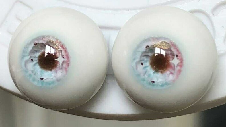 May include: Two realistic-looking glass eyes with a white sclera, a brown iris, and a blue and pink pattern. The eyes are painted with gold glitter.