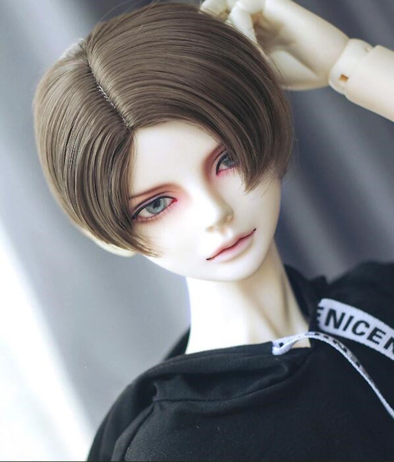 May include: A doll with brown hair styled in a short, side-swept cut. The doll is wearing a black hoodie with white text that reads "NICE".