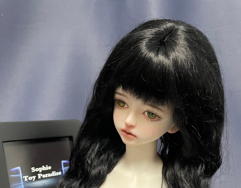 May include: A ball-jointed doll with pale skin, large amber-colored eyes, and long, wavy black hair with bangs. The doll has a neutral facial expression and is positioned next to a black sign displaying the text Sophie Toy Paradise in white.
