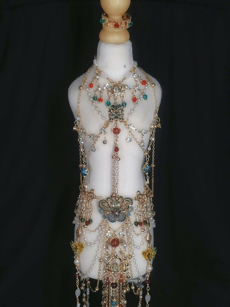 May include: A gold-tone body chain with multiple layers of beads and charms. The chain features a large, ornate pendant with blue and green enamel. The chain is decorated with red, green, and white beads.