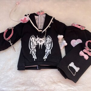 K&ouml;nnte beinhalten: Schwarzes und rosafarbenes Gothic-Kleidungsset mit einem schwarzen Sweatshirt mit einem wei&szlig;en Engelsfl&uuml;gel-Design mit Stacheldraht, einer schwarzen Hose mit einem rosafarbenen Herzen und einem wei&szlig;en Kreuz sowie einer rosafarbenen Halskette mit einem silbernen Anh&auml;nger.