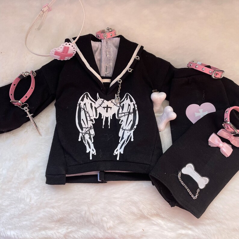 K&ouml;nnte beinhalten: Schwarzes und rosafarbenes Gothic-Kleidungsset mit einem schwarzen Sweatshirt mit einem wei&szlig;en Engelsfl&uuml;gel-Design mit Stacheldraht, einer schwarzen Hose mit einem rosafarbenen Herzen und einem wei&szlig;en Kreuz sowie einer rosafarbenen Halskette mit einem silbernen Anh&auml;nger.