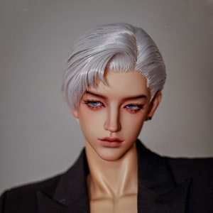 May include: A doll with short, silver-white hair styled in a side part. The doll has blue eyes and is wearing a black blazer.