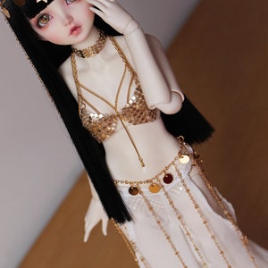 May include: A doll with long black hair wearing a gold sequined top and a white skirt with gold chains and coin accents.