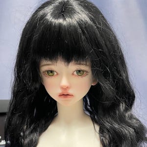 May include: A ball-jointed doll with pale skin, long wavy black hair with bangs, and light green eyes. The doll has a neutral facial expression with soft pink lips and defined eyelashes. A partial black label with the text Sophie Paradi is visible.