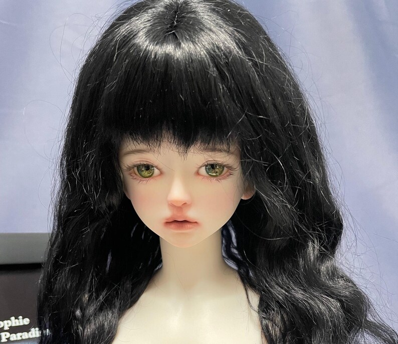 May include: A ball-jointed doll with pale skin, long wavy black hair with bangs, and light green eyes. The doll has a neutral facial expression with soft pink lips and defined eyelashes. A partial black label with the text Sophie Paradi is visible.