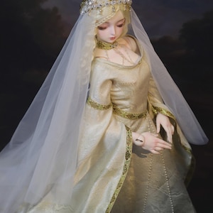 Puede incluir: Una muñeca articulada con un vestido largo de color oro pálido, mangas largas y anchas con bordes dorados. La muñeca lleva una gargantilla dorada a juego, un tocado con perlas y un largo velo blanco transparente sobre su cabello rubio.