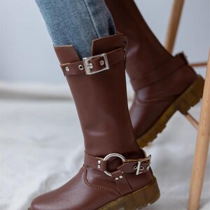May include: Brown leather knee-high boots with a buckle and a silver ring detail. The boots have a chunky heel and a zipper on the side.
