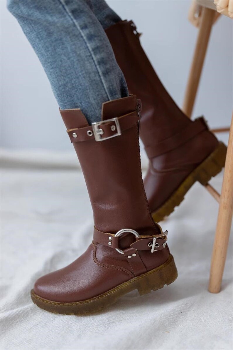 May include: Brown leather knee-high boots with a buckle and a silver ring detail. The boots have a chunky heel and a zipper on the side.