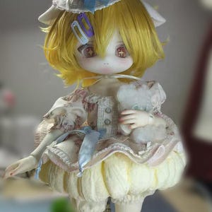 May include: A detailed doll with yellow hair, wearing a cream-colored dress with floral patterns and a matching hat topped with a red cherry. The doll has large eyes and is holding a small stuffed animal.