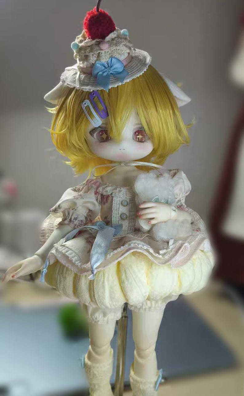 May include: A detailed doll with yellow hair, wearing a cream-colored dress with floral patterns and a matching hat topped with a red cherry. The doll has large eyes and is holding a small stuffed animal.