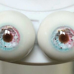 May include: Two white doll eyes with blue, pink, and brown irises and gold glitter accents.