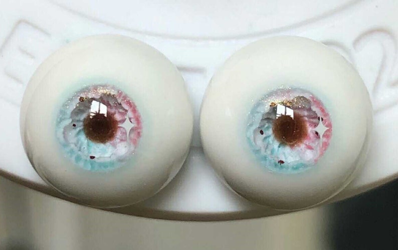 May include: Two white doll eyes with blue, pink, and brown irises and gold glitter accents.