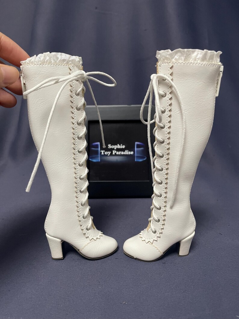 Pode incluir: Um par de botas de boneca brancas com design de cadar&ccedil;o e saltos pequenos. As botas t&ecirc;m uma borda com babados na parte superior e costuras decorativas nas laterais. S&atilde;o exibidas em frente a uma caixa preta com o texto "Sophie Toy Paradise".
